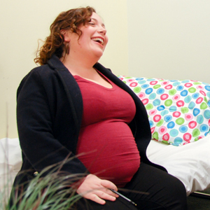 Pregnant person seated indoors with face blurred, wearing a red top and black pants, holding a pen, with a bed featuring polka dot bedding in the background.